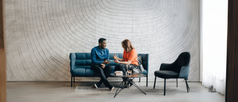 Two colleagues sitting on a sofa in a modern office lounge, having a friendly conversation with laptops and notebooks on the table.